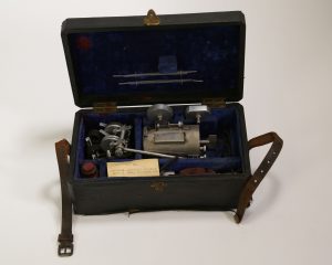 Photograph of a Mackenzie Polygraph. The case containing the object is open to show the parts of the object, which are not assembled.