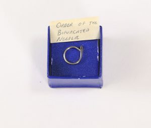 Photo of a lapel pin in a blue box. The pin is made from a bifurcated needle that has been twisted into a circle.