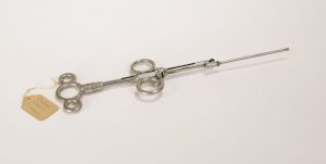 Photograph of a tonsil ecraseur. The object is made of metal and is shaped in a straight line with finger loops at the bottom and center of the object. There is no wire.
