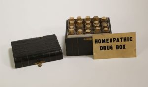 Photograph of a small chest containing small corked bottles. The chest is black. The lid is not attached and is sitting next to the part containing the bottles. A card that reads "HOMEOPATHIC DRUG BOX" is propped up against the chest.